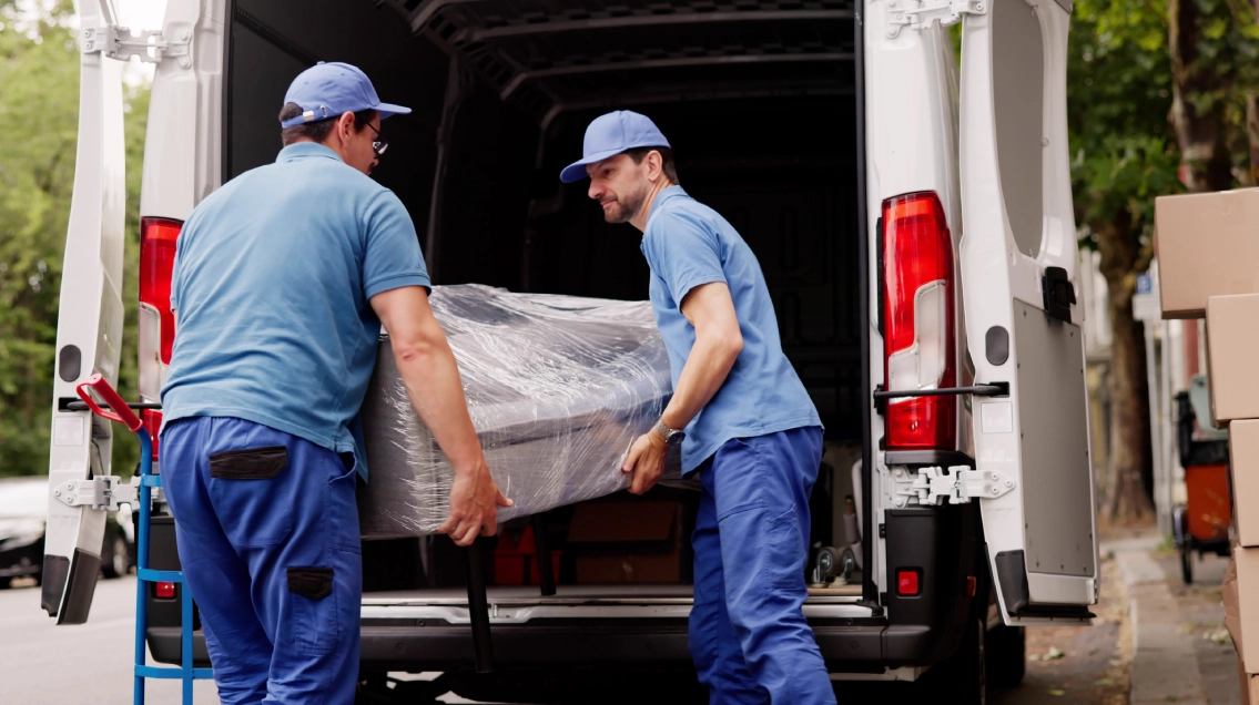 Two movers carrying a wrapped piece of furniture in a truck.